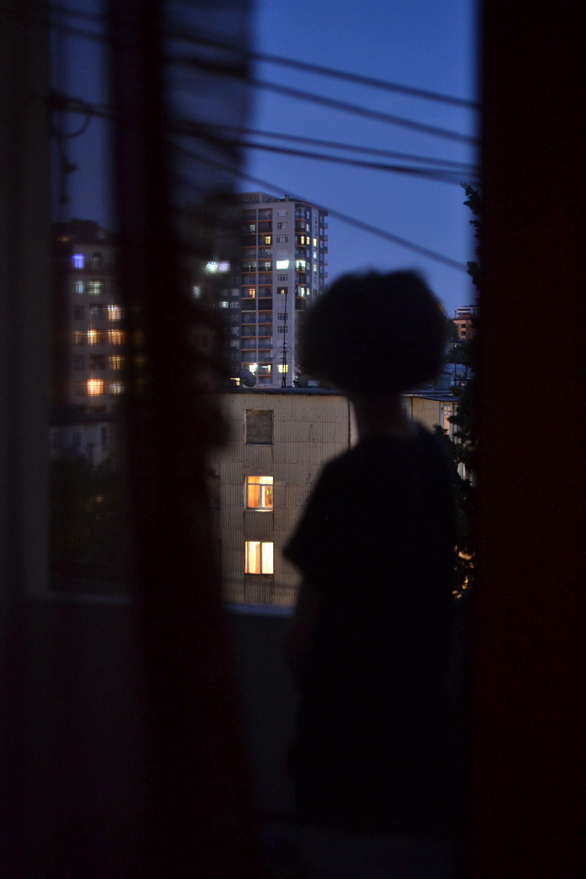 A woman at a window at night, looking across at the lit windows of another apartment building.