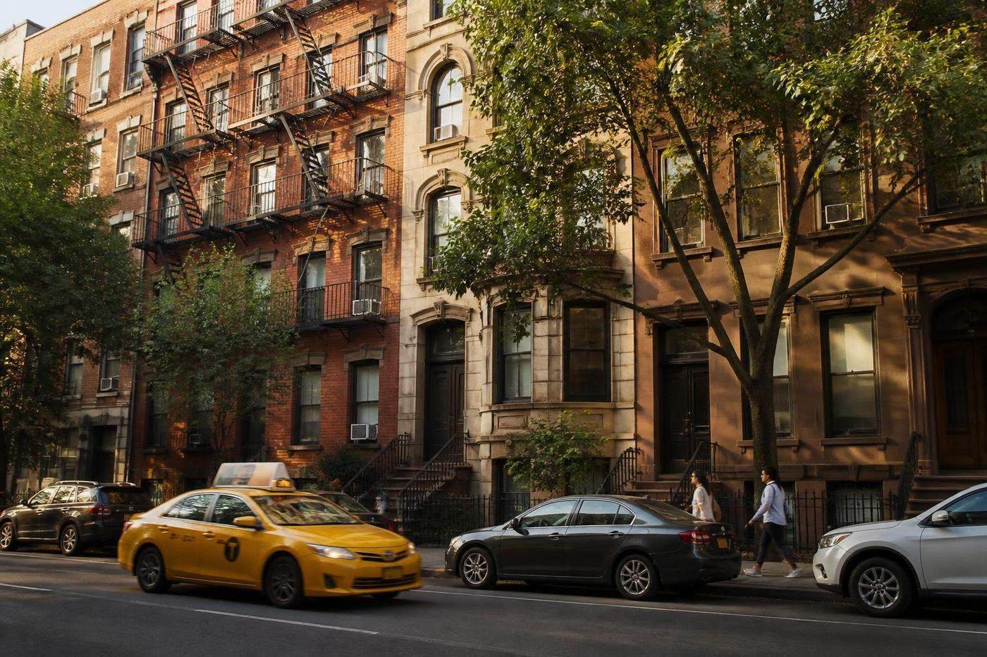 New York City apartment buildings along a tree-lined street with window air conditioners and traffic.