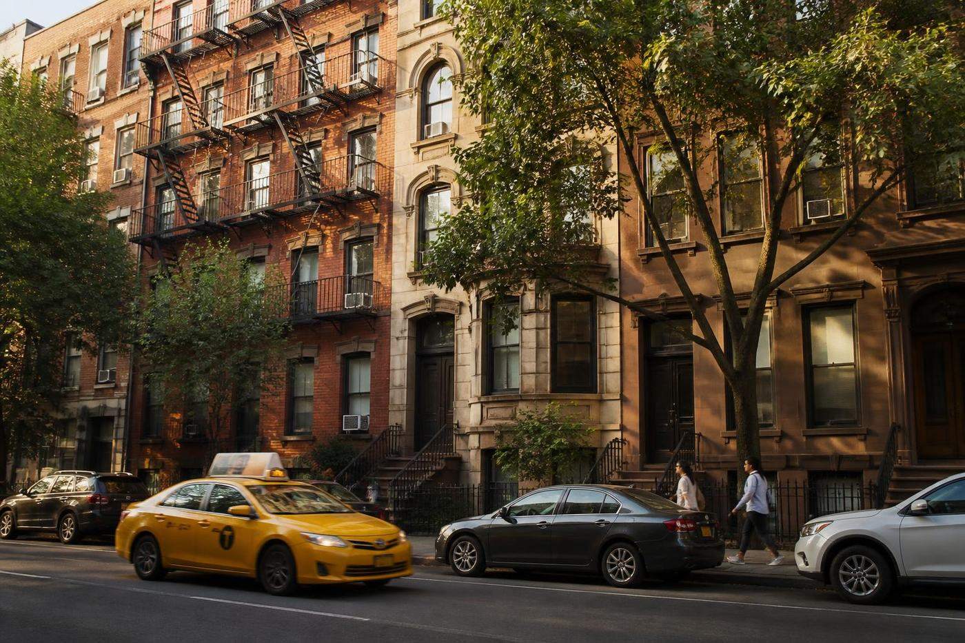New York City apartment buildings along a tree-lined street with window air conditioners and traffic.